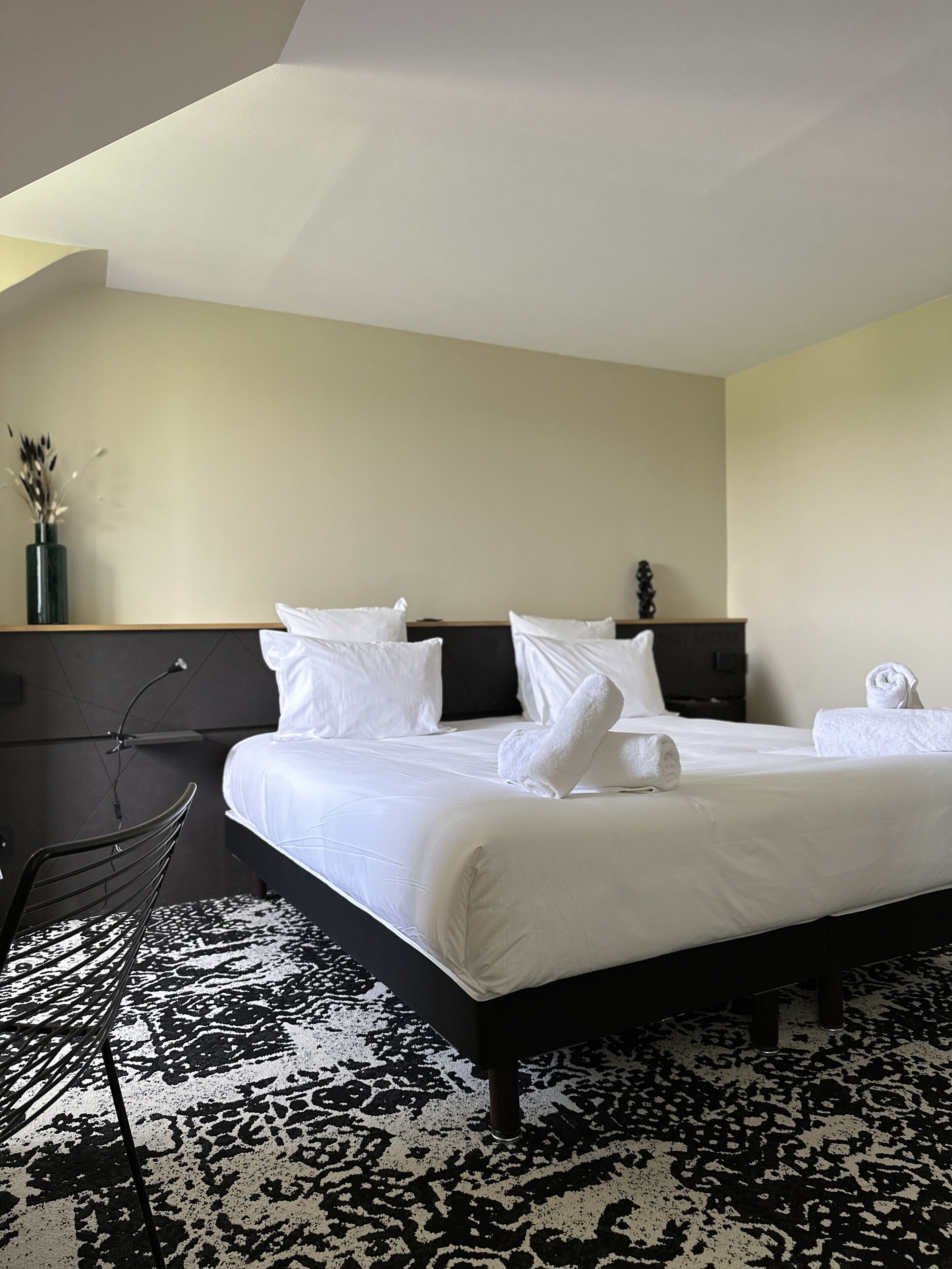 A hotel room with a large bed, a black metal chair, a vase with dried flowers, and a black and white patterned rug.