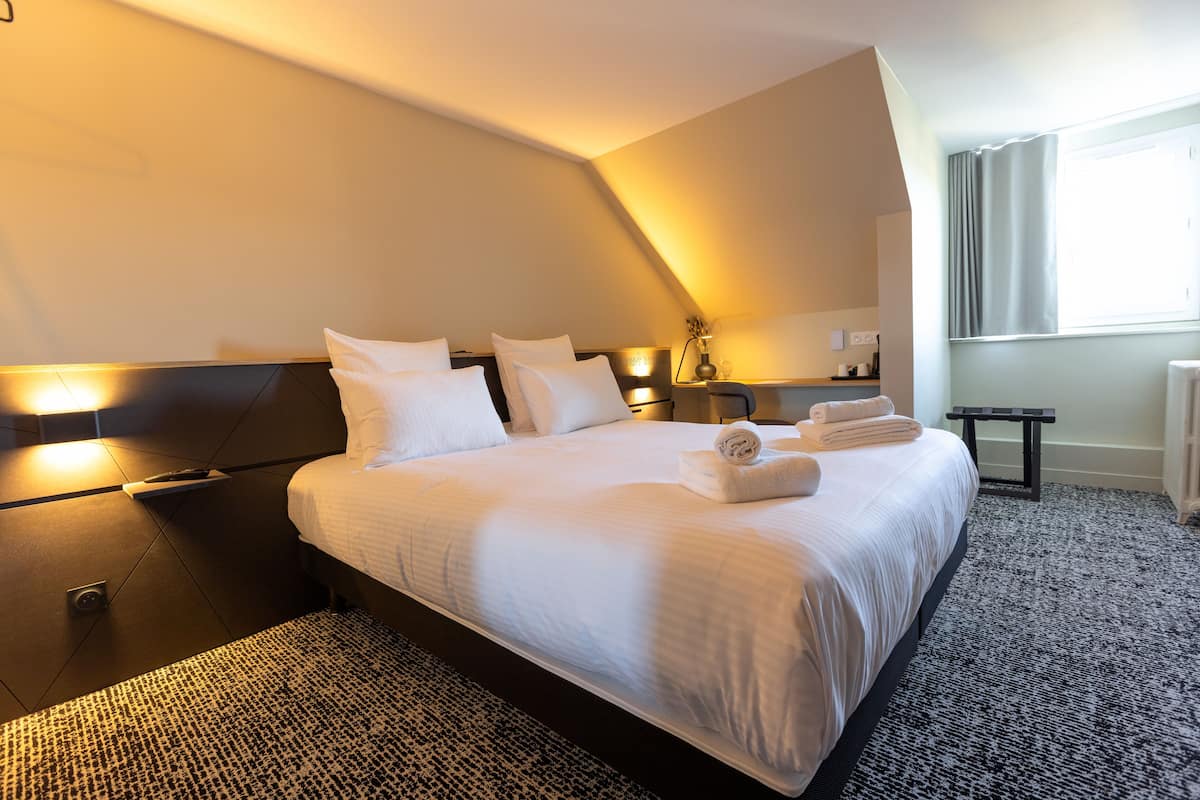 Hotellihuone, jossa on suuri sänky, yöpöytä, lamppu ja ikkuna, jossa on verhot.