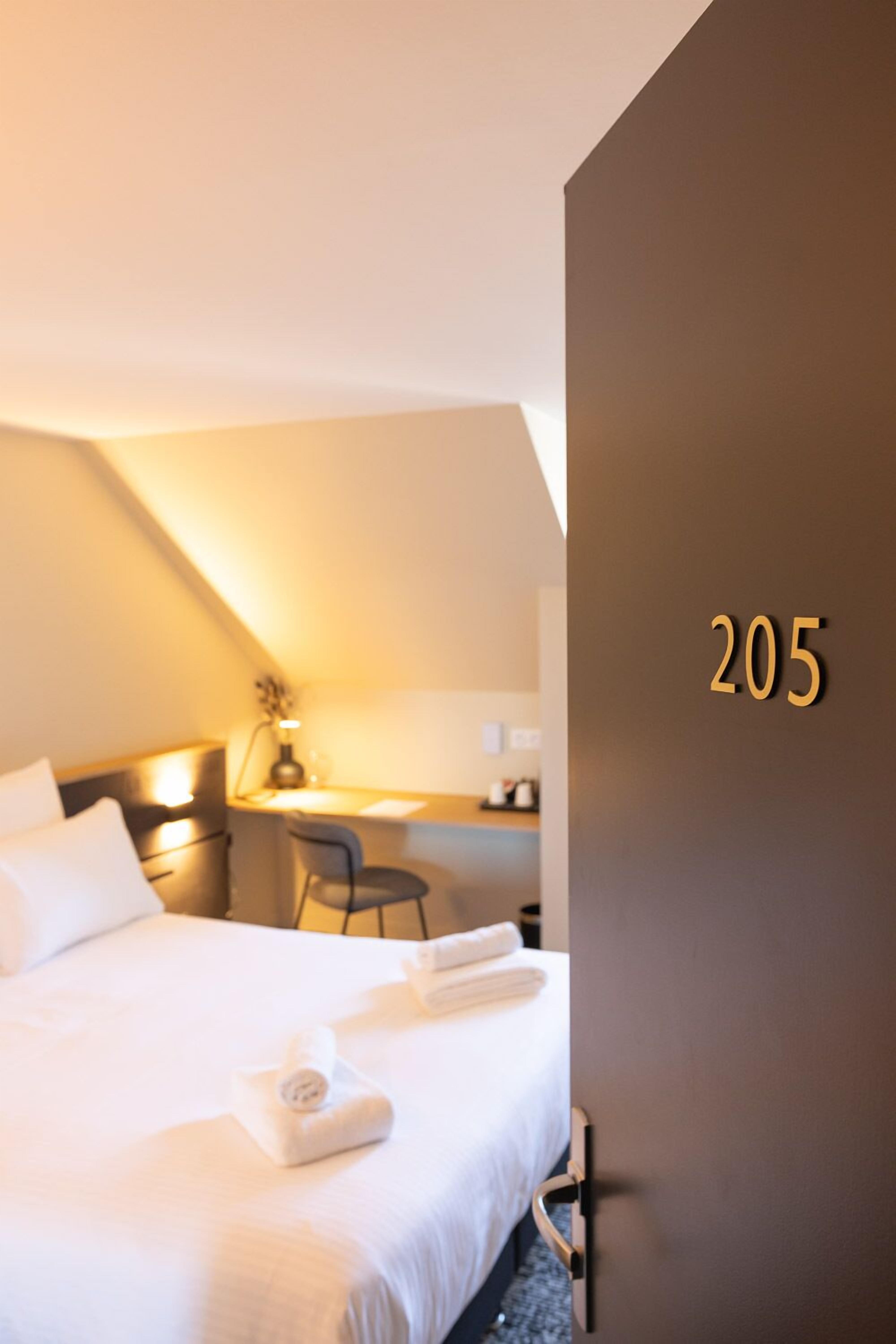 A hotel room with a neatly made bed, a desk with a chair, and a wall with the number 205.