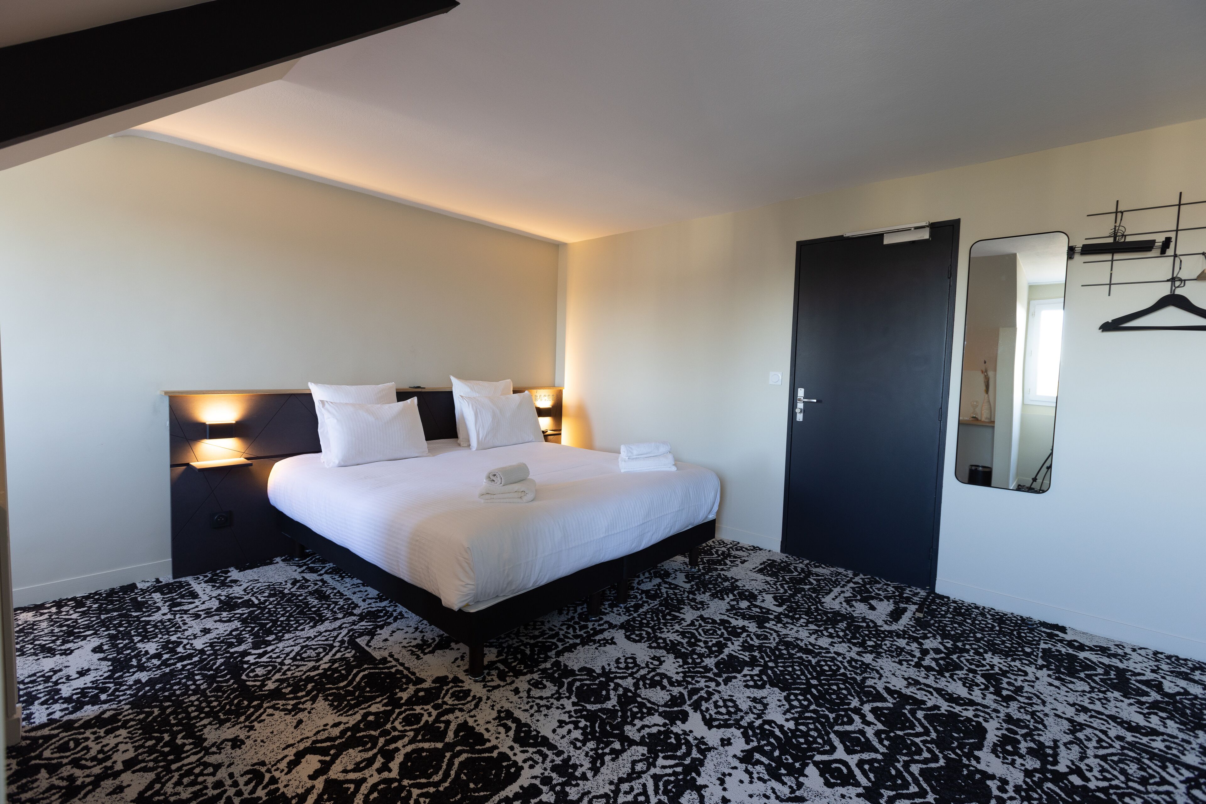 Une chambre d’hôtel avec un grand lit, une table de chevet avec une lampe, une porte noire et un miroir fixé au mur.