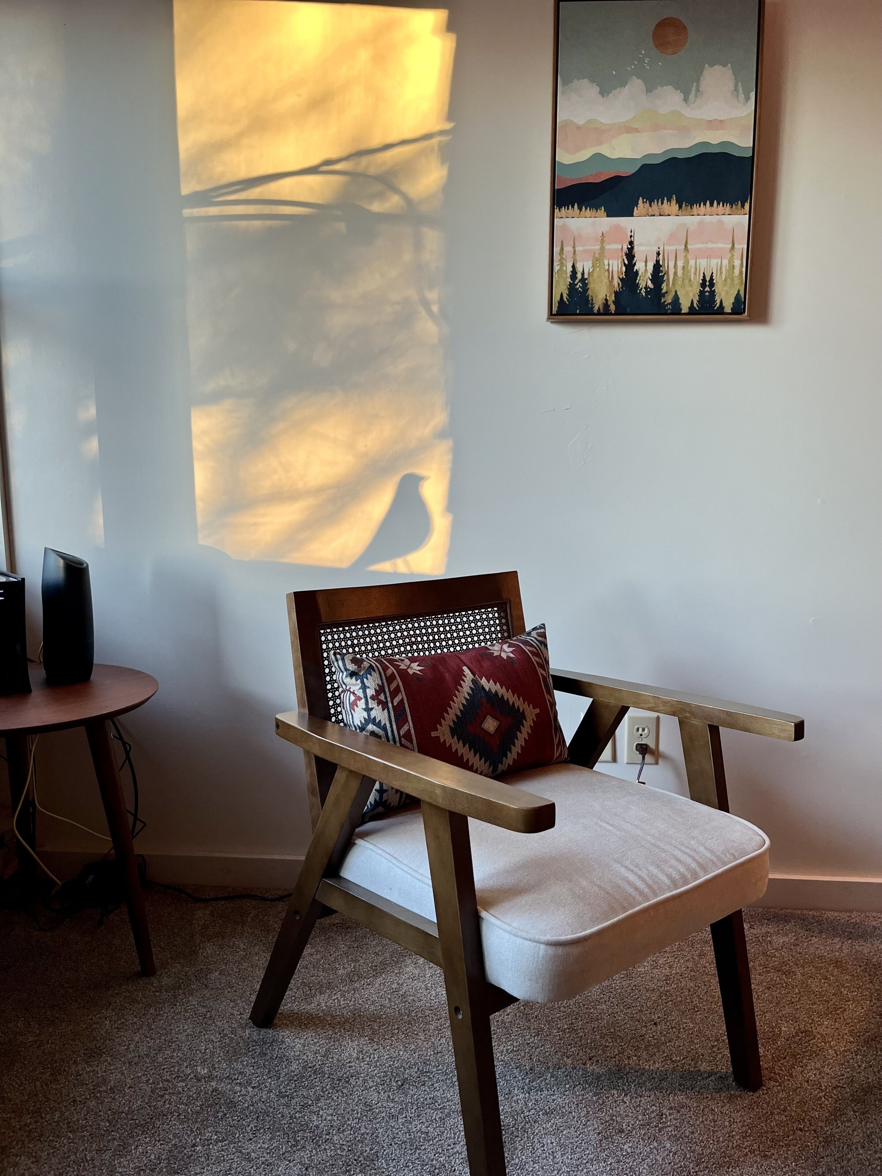 Ein Zimmer mit einem Holzstuhl, einem Kissen, einem kleinen runden Tisch und einem an der Wand hängenden Gemälde.