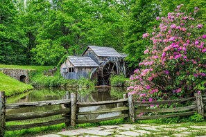 Eine rustikale Wassermühle mit Holzkonstruktion und einem großen Wasserrad, umgeben von üppiger Vegetation und einer kleinen Brücke.