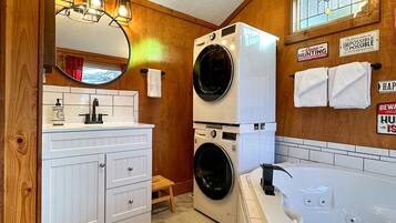 Ein kompaktes, gut beleuchtetes Badezimmer mit einer Waschtrockner-Kombination für zwei Personen, einem kleinen Holzschemel, einem großen Spiegel und einer Badewanne.