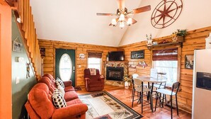 Un interior acogedor de cabaña con chimenea, paredes de madera, zona de comedor y un área de estar confortable con sofá y sillones.