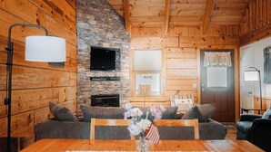 Un interior de cabaña acogedor con chimenea de piedra, paredes de madera, una mesa de comedor con mantel de encaje y un sofá con cojines.
