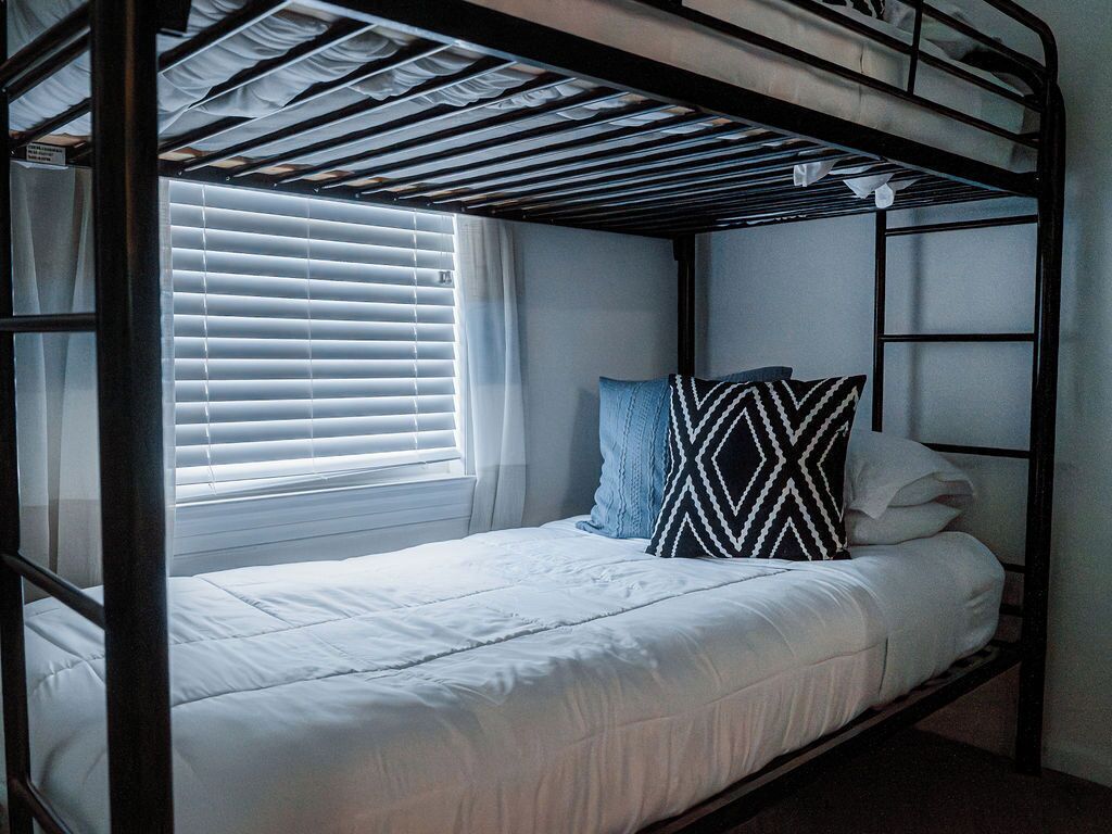 A bunk bed with a white mattress and a patterned pillow, a window with blinds, and a metal frame.