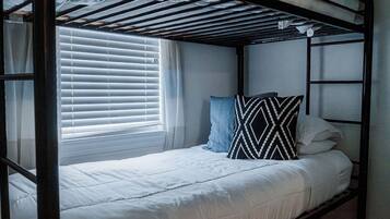 A bunk bed with a white mattress and a patterned pillow, a window with blinds, and a metal frame.