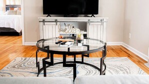 A modern living room with a round glass coffee table, a flat-screen TV mounted above a white cabinet, and a bed with a white headboard in the background. - "Mountain Escape" - Majestic Mountain Sunrises Near Blue Ridge Parkway & I-77 (Fancy Gap)