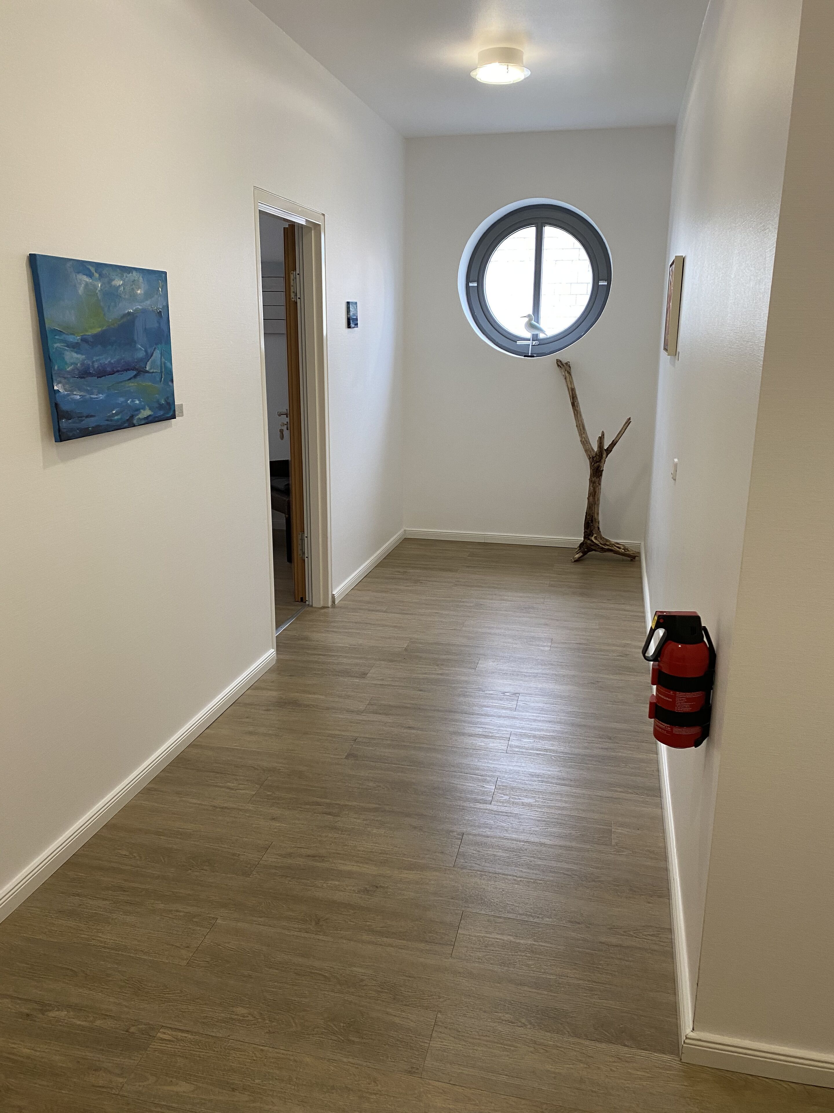 Un pasillo con piso de madera, un ventanal redondo, un extintor de incendios montado en la pared y un cuadro.