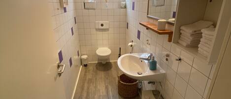 A modern bathroom with a toilet, sink, and bathtub. There is a window above the toilet and a shelf with towels.