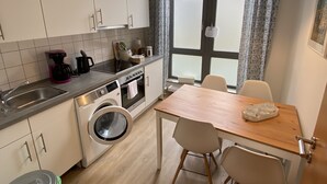 A compact kitchen with white cabinets, a washing machine, a coffee maker, and a dining table with chairs. - Gästehaus Lavendel (Flensburg)