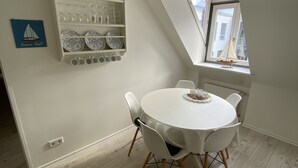 A dining area with a round table, white chairs, a white wall, a shelf with dishes, and a small sailboat model by a window. - Gästehaus Lavendel (Flensburg)