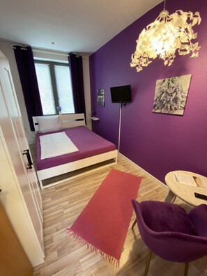 Un dormitorio con cama, televisor, pared morada y ventana con cortinas.