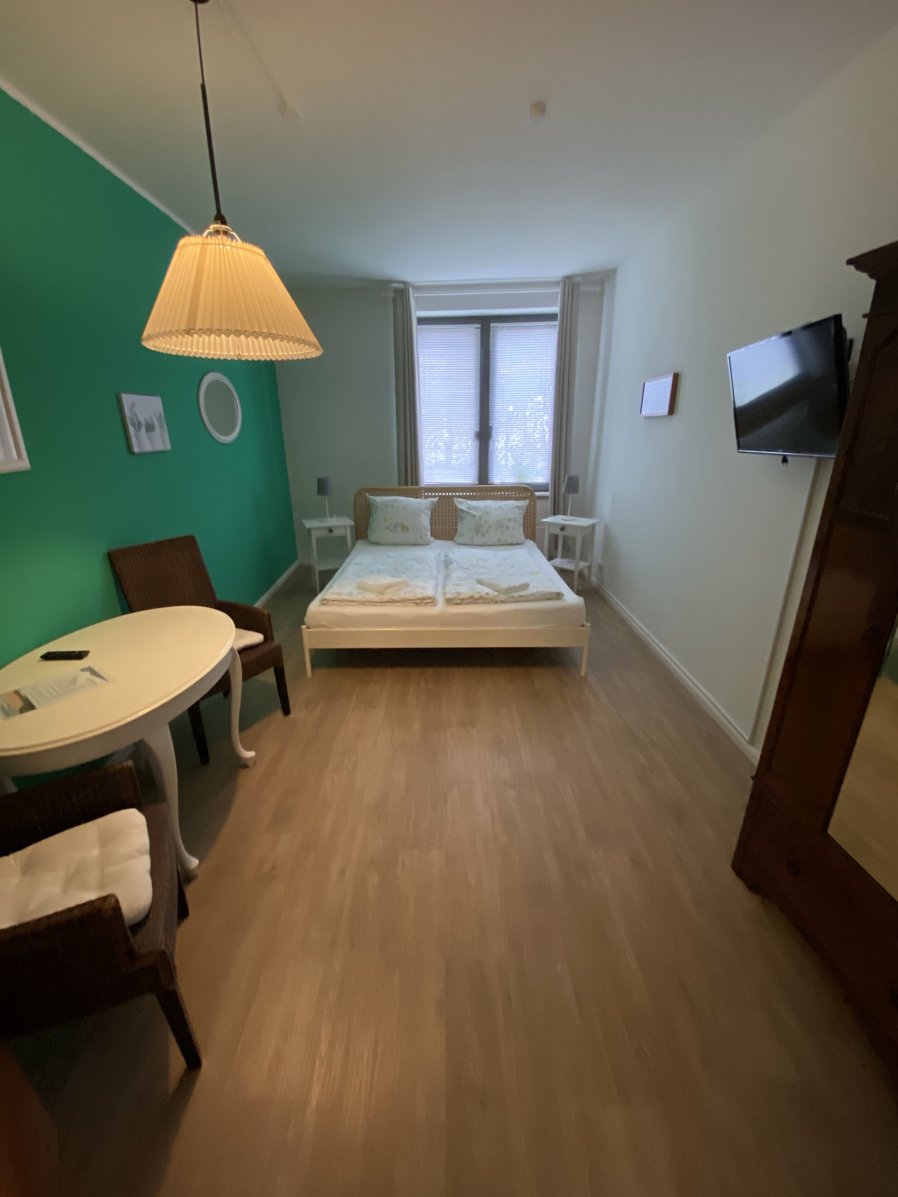 A bedroom with a bed, a desk, and a chair. There is a window with blinds, a TV on the wall, and a hanging lamp.