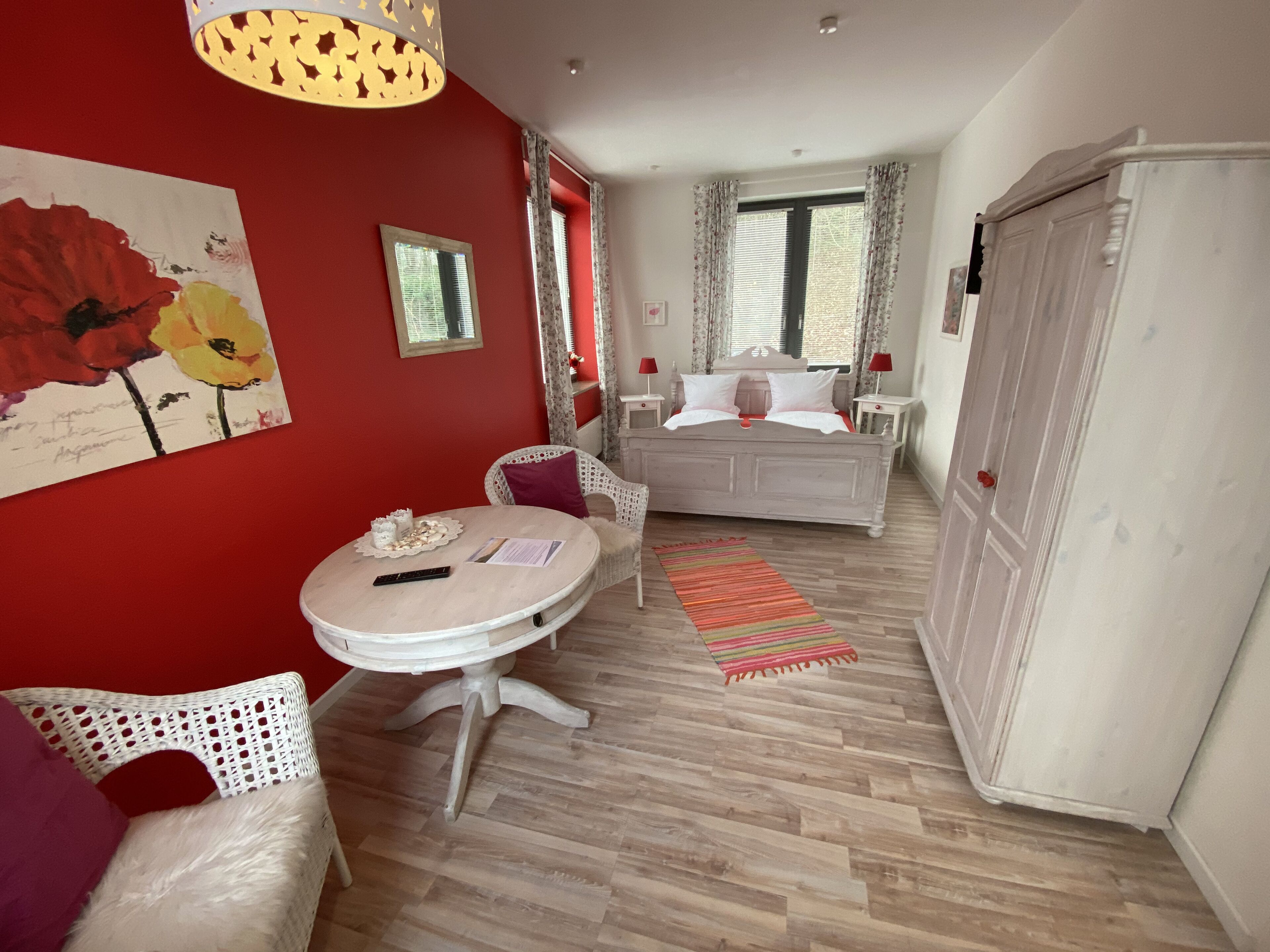 A room with a bed, a round table with a remote control, a white wardrobe, and a red wall with a floral painting.