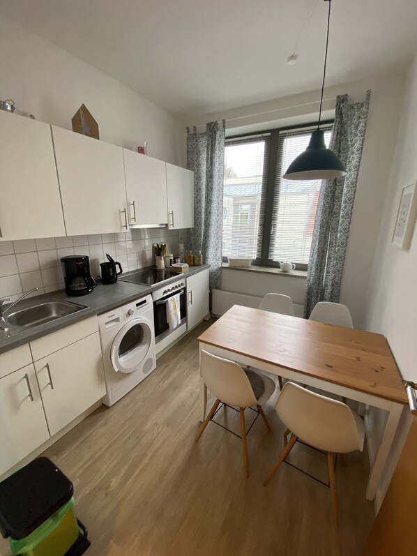 Una cocina compacta con armarios blancos, horno empotrado, lavadora y un comedor con mesa y sillas de madera.