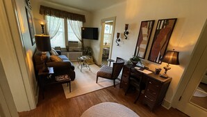 Een gezellige woonkamer met een bank, een salontafel, een bureau en een stoel. Er is een televisie aan de muur bevestigd en een raam met gordijnen.