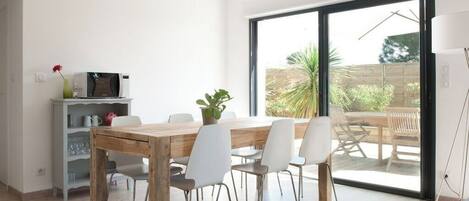 A modern dining area with a wooden table and white chairs, a small shelf with kitchen items, and a view through a large sliding glass door to an outdoor patio.
