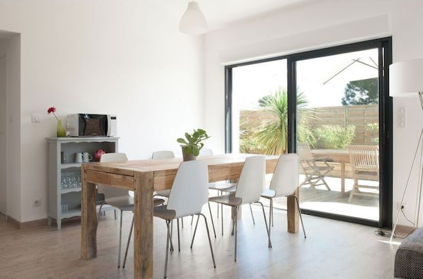 Un espace repas moderne avec une table en bois et des chaises blanches, une petite étagère contenant des ustensiles de cuisine, et une vue sur une terrasse extérieure, accessible par une grande porte-fenêtre coulissante.