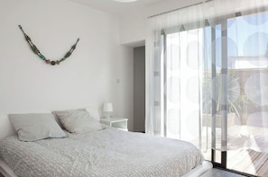 Une chambre à coucher avec un lit, une table de chevet, une lampe et une vue sur l’extérieur à travers des rideaux légers.