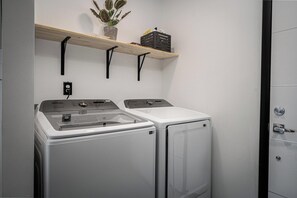 A laundry area with two white washing machines, a wooden shelf with a potted plant and storage boxes, and a door with a handle.