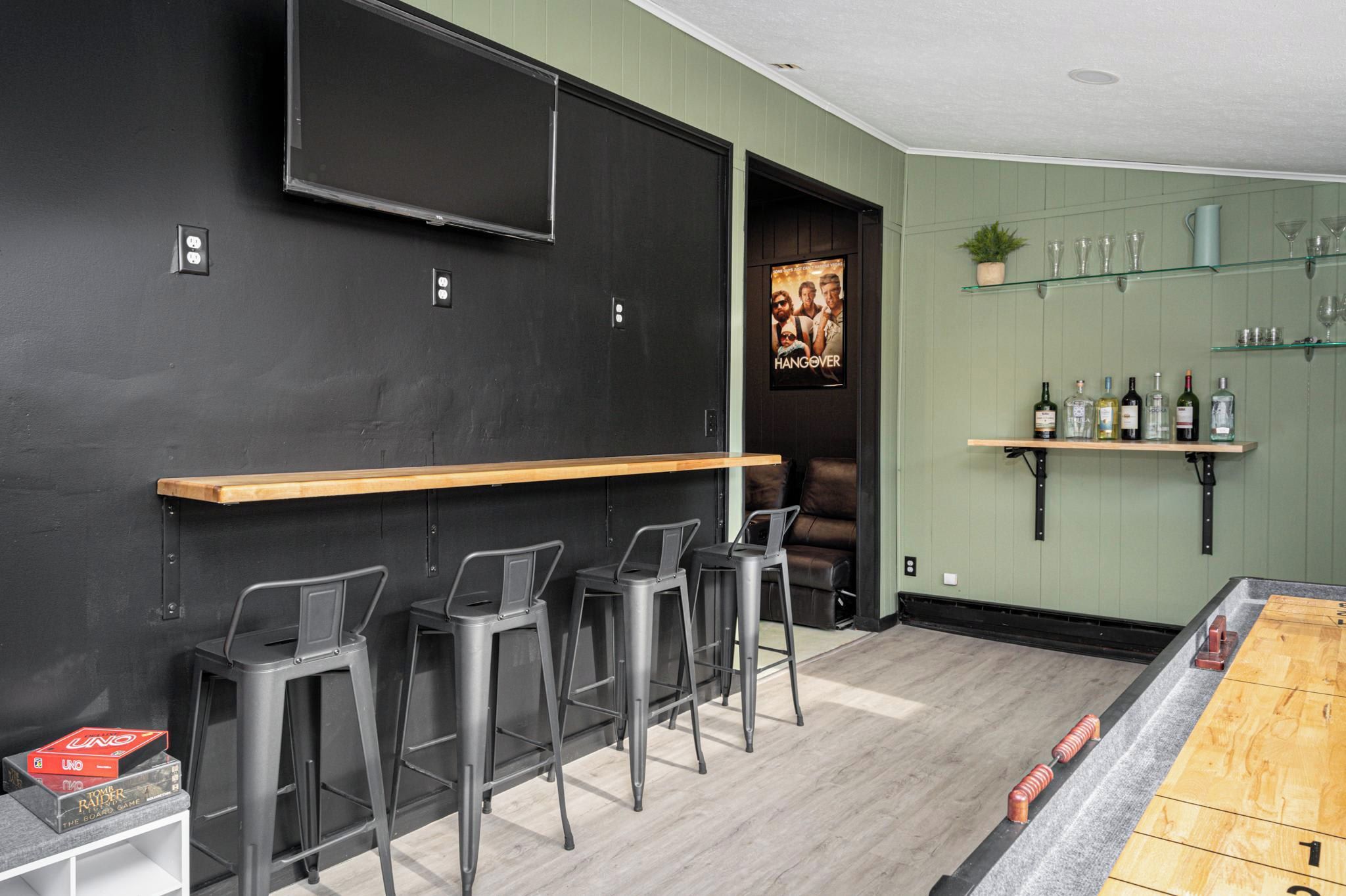 A bar area with a long wooden counter, black walls, and three metal bar stools.