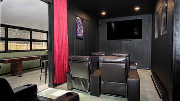 Una sala de cine en casa con un televisor grande, sillones de cuero reclinables, una cortina roja y un banco de madera.