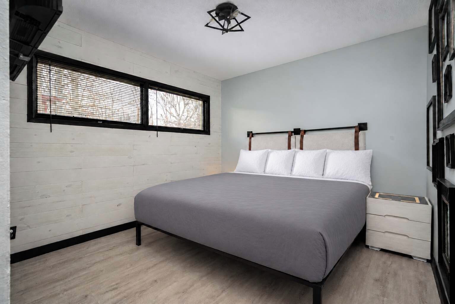 A bedroom with a bed, a nightstand, a window with blinds, and a ceiling light fixture.
