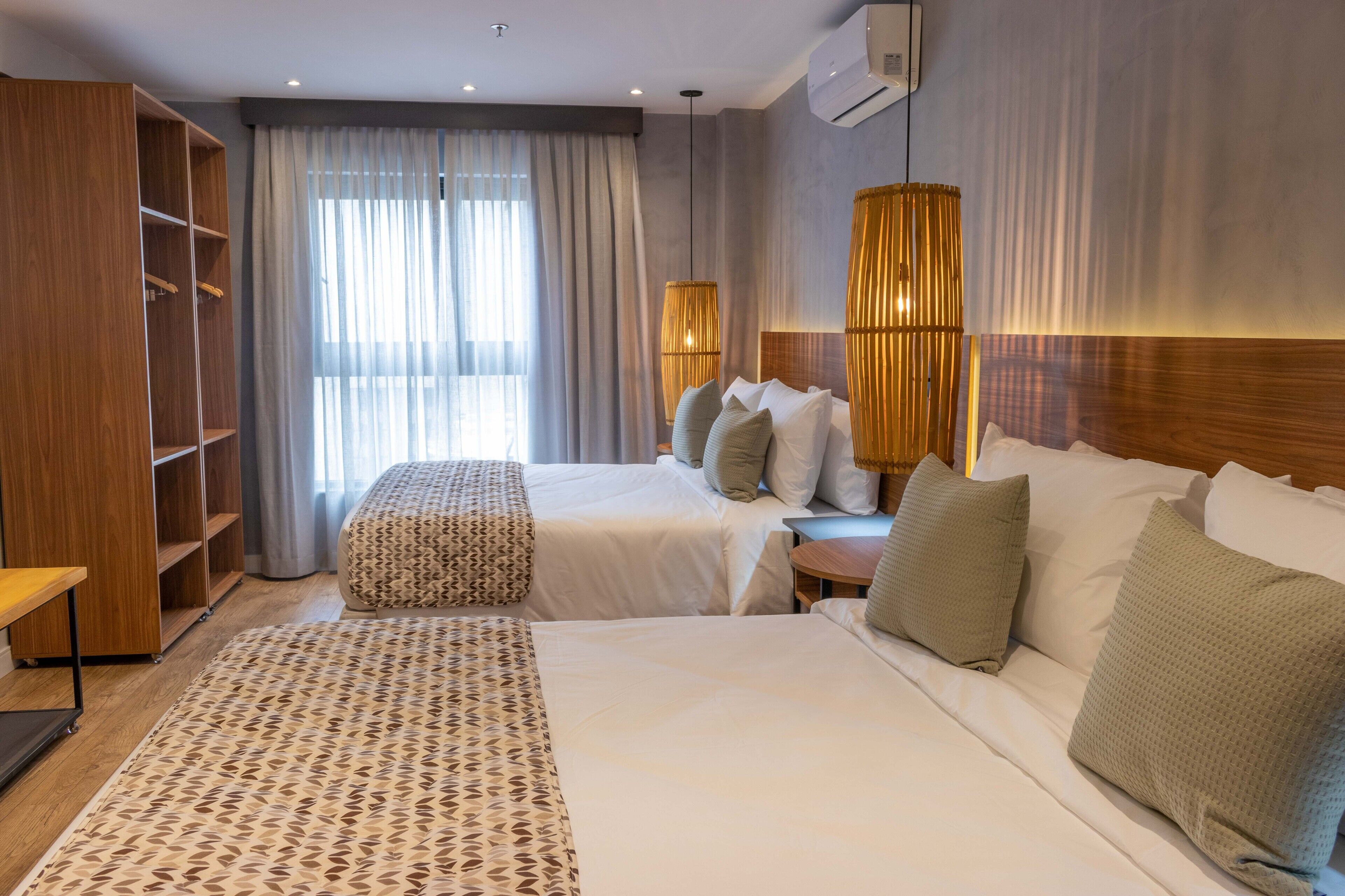 A hotel room with two beds, a wooden wardrobe, a patterned rug, and a large window with curtains.