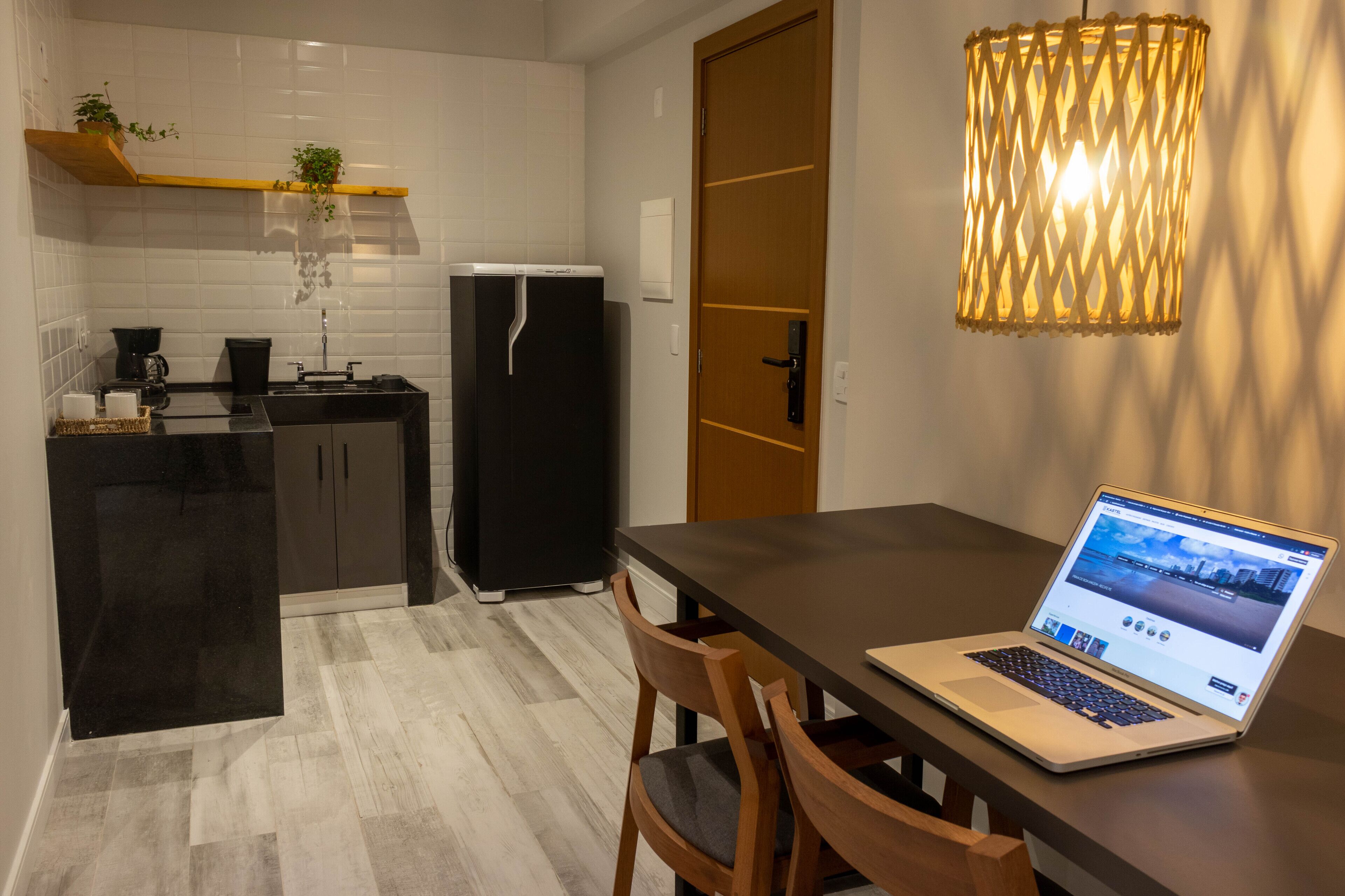 A modern kitchen with a black refrigerator, a coffee machine, and a wooden shelf with plants. A laptop is on a table with chairs.