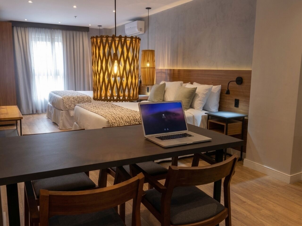 A modern hotel room with a wooden dining table, a laptop, and a bed with a headboard.