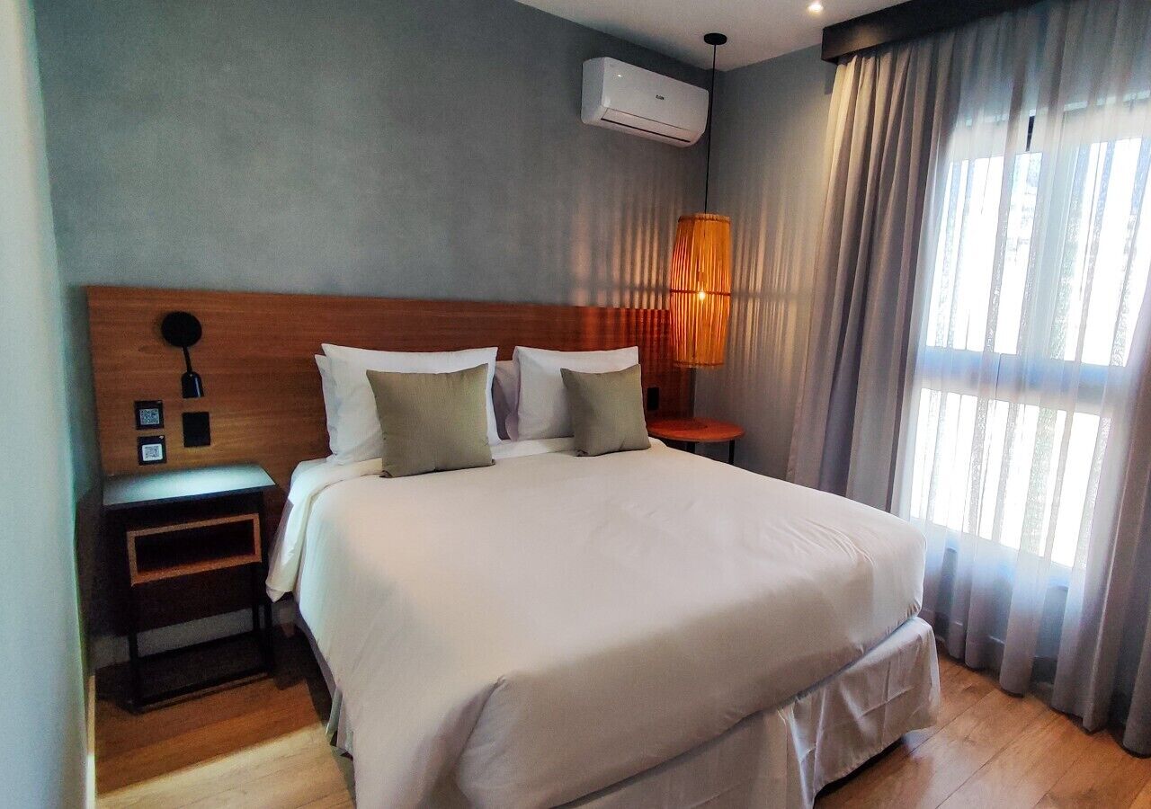 A neatly made bed with a wooden headboard, a bedside table with a lamp, and a window with curtains.