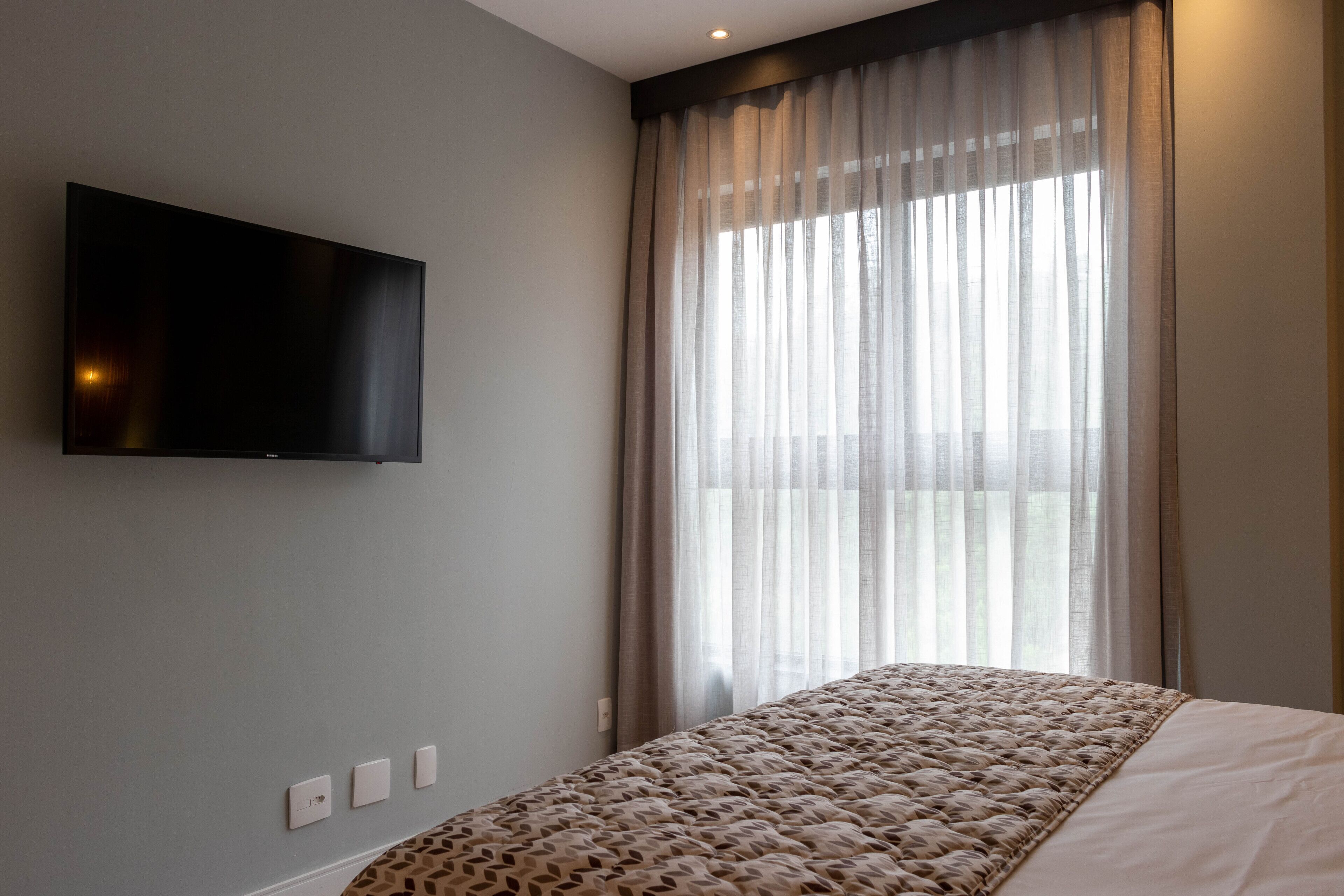 A modern hotel room with a large bed, a mounted TV, and a large window with sheer curtains.