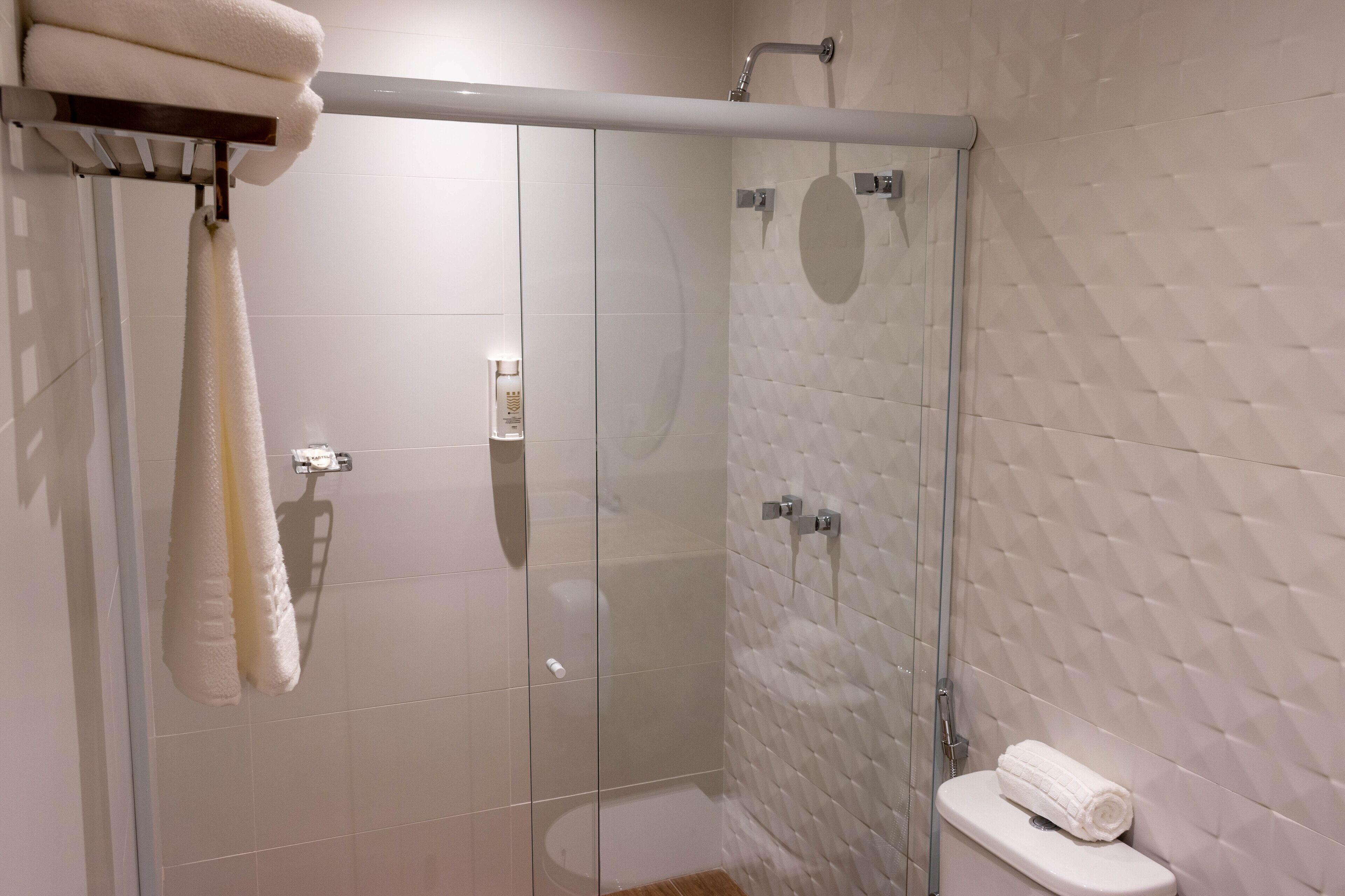 Um chuveiro com divisória de vidro, parede revestida de azulejos brancos e um vaso sanitário com toalha branca.