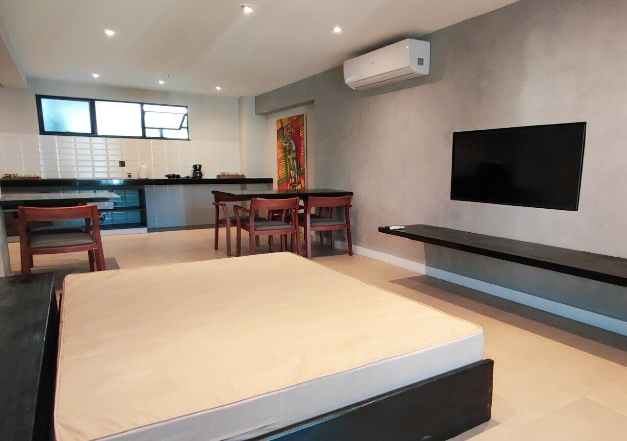 A modern living room with a large bed, a TV mounted on the wall, and a kitchen area in the background.
