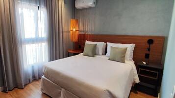 A neatly arranged hotel room with a large bed, a bedside table, a lamp, and an air conditioning unit.