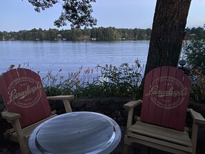 Duas cadeiras Adirondack com uma mesa de metal na frente, localizadas perto de um lago, com uma árvore e um edifício ao fundo.