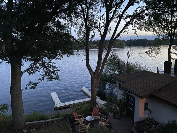 Uma propriedade à beira de um lago com um cais que se estende para a água, rodeada por árvores e um pequeno edifício com telhado inclinado.