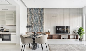 A modern living room with a marble dining table, grey upholstered chairs, a flat-screen TV, and a large abstract painting on the wall. - WelHome - Deluxe Apartment With Direct Access to the Beach (Dubai)