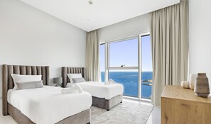 Two large beds with grey headboards and white bedding, a sliding glass door to a balcony with sea views.