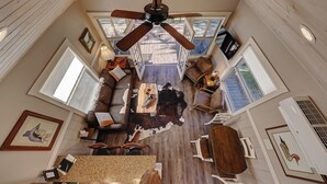 A spacious living area with a ceiling fan, a large wooden table, leather sofas, and a view of the deck through large windows.