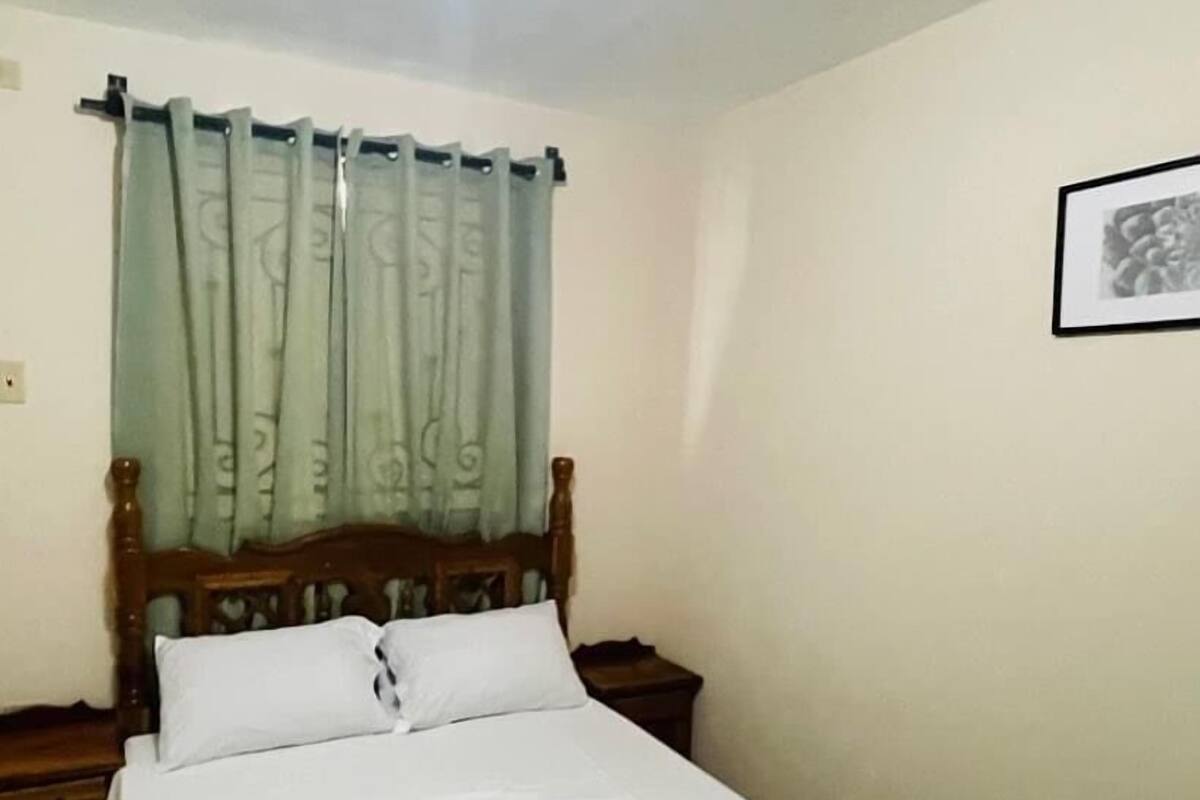 Ein ordentlich bezogenes Bett mit grüner Bettdecke in einem schlichten Zimmer, mit einem Holz-Kopfteil und einem Nachttisch.