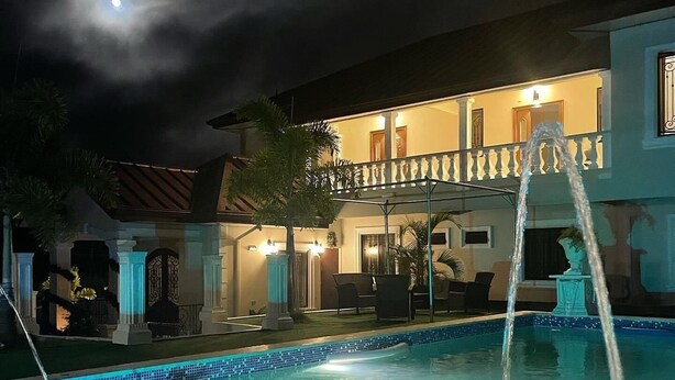 Une piscine avec une fontaine, un balcon agrémenté de mobilier d’extérieur et un bâtiment dont les fenêtres sont illuminées la nuit.