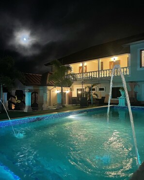 A swimming pool with a water fountain, a balcony with outdoor furniture, and a building with illuminated windows at night. - Vintage Oaks Hideaway  (Princes Town)