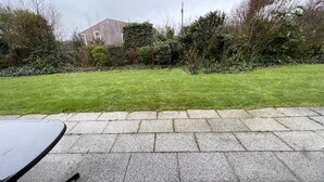 A paved patio area with a table and chairs, surrounded by a well-maintained lawn and dense shrubbery.