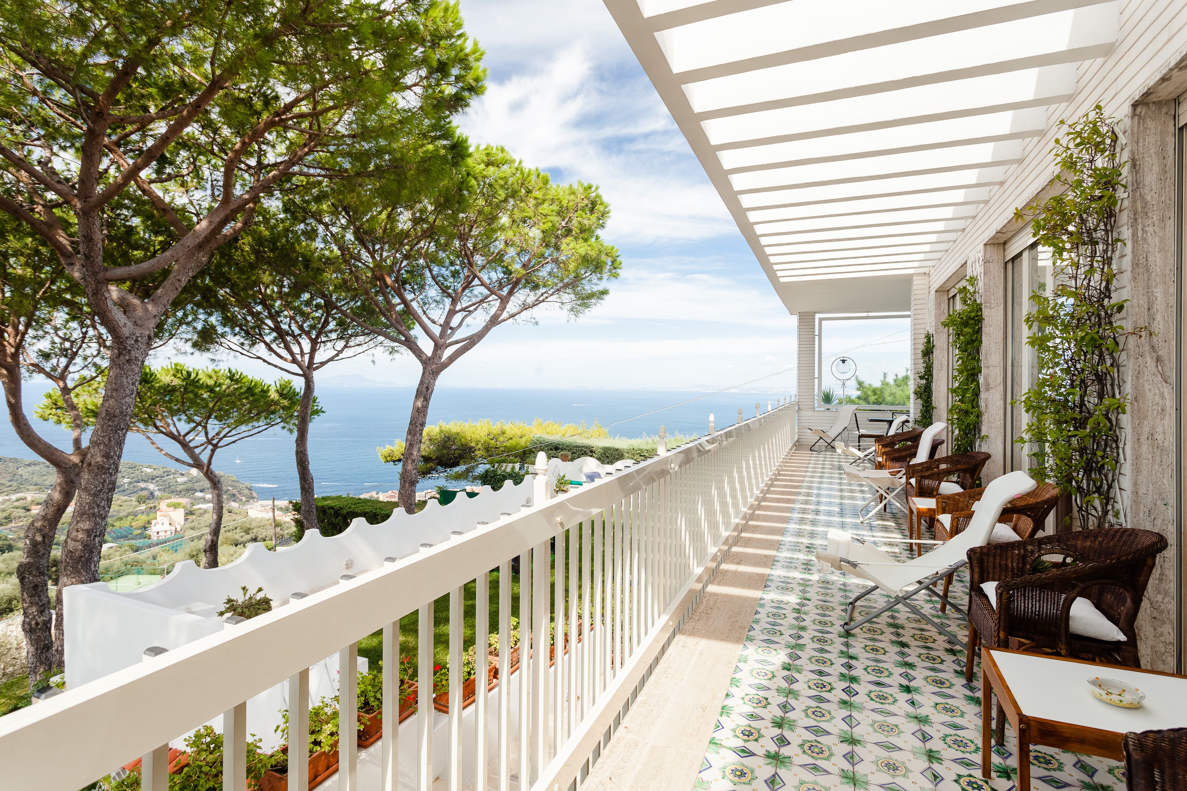 A balcony with wicker chairs and a tiled floor, overlooking a coastal landscape with trees and a view of the sea.