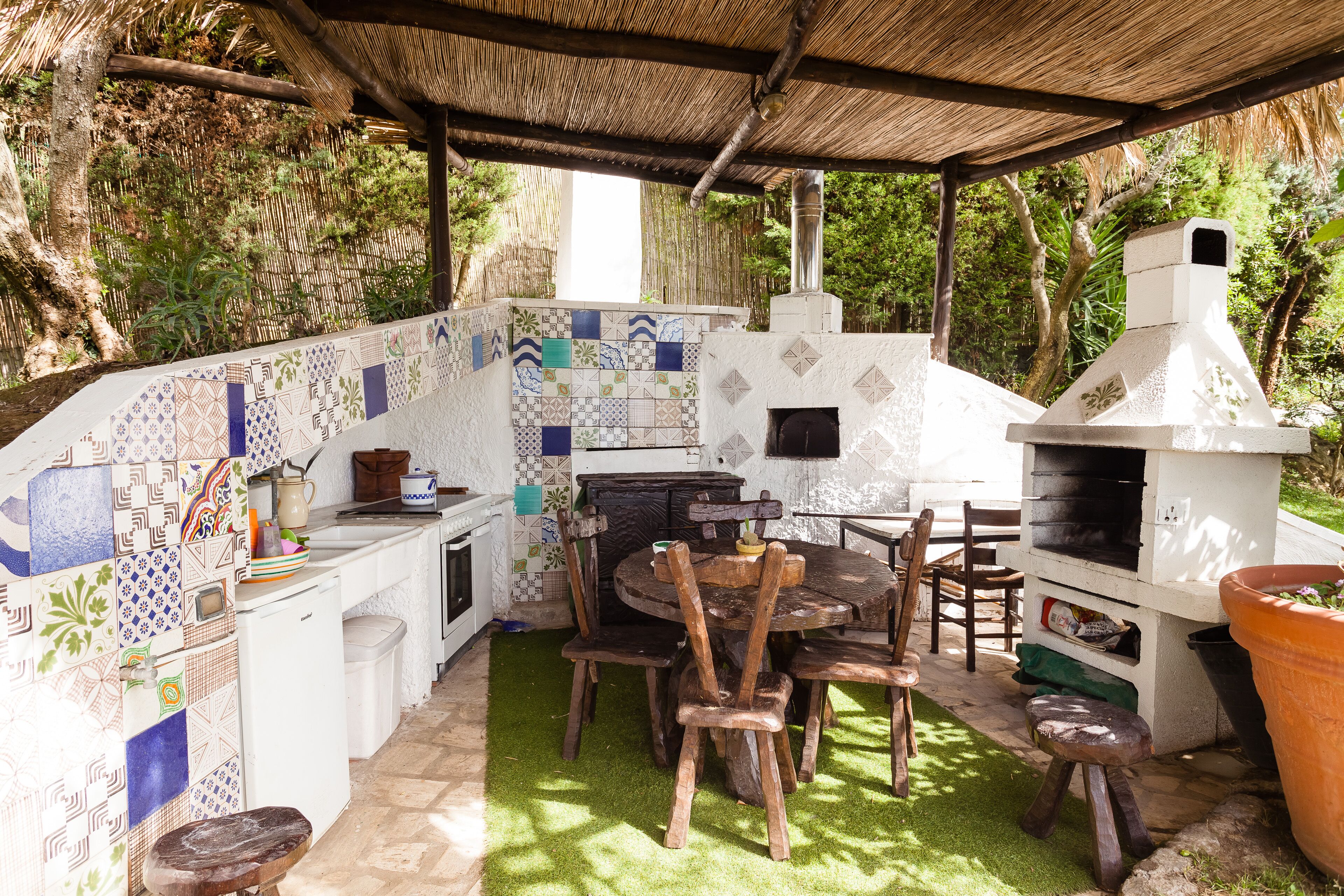 Une cuisine extérieure équipée d’un barbecue, d’une table et de chaises, le tout entouré d’un mur recouvert de carreaux et de verdure.