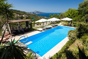 A rectangular pool with a clear blue water surface, surrounded by lounge chairs and umbrellas, set against a backdrop of lush greenery and a distant view of the sea and mountains. - Villa Soleil de Capri (massa lubrense)