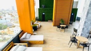 A modern rooftop lounge with wooden beds, a green wall, and a view of the cityscape. - Condos Ferrara (Mazatlán)