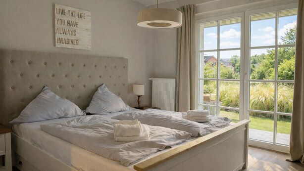 A bedroom with a large bed, a headboard, a bedside table with a lamp, and a window with a view of greenery. - Beautiful apartment with large south-facing terrace (Sankt Peter-Ording)